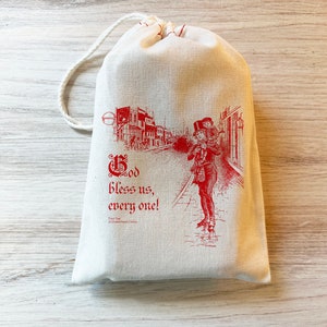 K&ouml;nnte beinhalten: Ein cremefarbener Stoffbeutel mit Kordelzug und einem roten Aufdruck, der eine Person zeigt, die ein Kind auf den Schultern durch eine Stra&szlig;e tr&auml;gt. Der Text lautet "God bless us, every one!" sowie "Tiny Tim, A Christmas Carol" in roter Farbe.