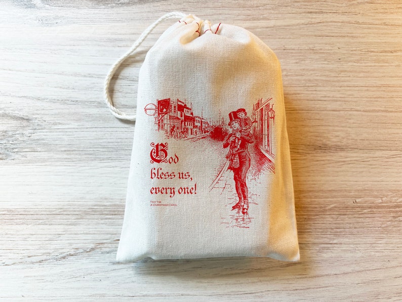 K&ouml;nnte beinhalten: Ein cremefarbener Stoffbeutel mit Kordelzug und einem roten Aufdruck, der eine Person zeigt, die ein Kind auf den Schultern durch eine Stra&szlig;e tr&auml;gt. Der Text lautet "God bless us, every one!" sowie "Tiny Tim, A Christmas Carol" in roter Farbe.