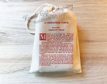 May include: A cream-colored drawstring gift bag featuring red printed text from A Christmas Carol, Stave One: Marley's Ghost. The bag includes a decorative initial M and the text Kport Gift Company Handmade Gift Bags at the top.