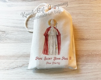 May include: A cream-colored fabric drawstring gift bag featuring a watercolor illustration of Pope Saint John Paul II in white and red vestments. The text "POPE SAINT JOHN PAUL II" and "Pray For Us" is printed in red below the central figure.