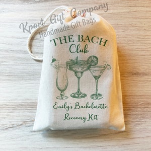 May include: A cream-colored drawstring fabric bag featuring a green graphic of three cocktail glasses. The text reads THE BACH Club, Emily's Bachelorette Recovery Kit, and Kport Gift Company Handmade Gift Bags in green ink.