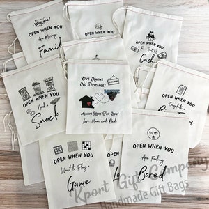 May include: Ten white cotton drawstring bags with black text and images. The bags have different sayings on them, such as "Open when you are missing family", "Open when you need a snack", "Open when you want to play a game", "Open when you are feeling bored", "Open when you run out of cash", "Open when you complete a ...", and "Love knows no distance! Always here for you! Love, Mom and Dad".