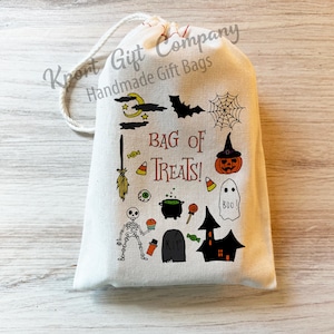 May include: A cream-colored Halloween-themed gift bag with a drawstring closure. The bag is decorated with various Halloween icons, including a bat, spiderweb, pumpkin, ghost, and the text "BAG OF TREATS!"