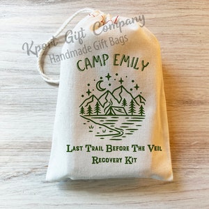 May include: A cream-colored drawstring fabric gift bag featuring a green graphic print of a mountain landscape with a tent, trees, a crescent moon, and stars. The text reads CAMP EMILY, LAST TRAIL BEFORE THE VEIL, and RECOVERY KIT in green ink.