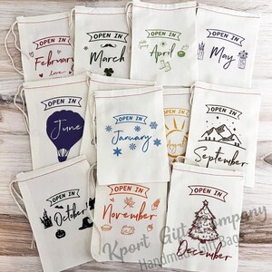 May include: Twelve white fabric drawstring bags with different months of the year printed on them. Each bag has a different design and color scheme. The bags are arranged in three rows of four. The text on the bags reads: "Open in February", "Open in March", "Open in April", "Open in May", "Open in June", "Open in January", "Open in August", "Open in September", "Open in October", "Open in November", "Open in December".