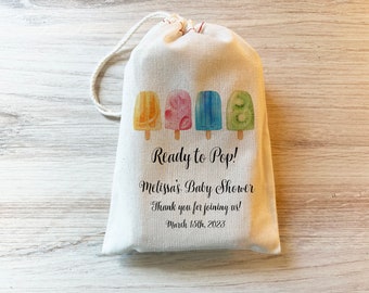 May include: A white fabric bag with a drawstring closure. The bag has a colorful illustration of popsicles and the text "Ready to Pop! Melissa's Baby Shower Thank you for joining us! March 15th, 2023".