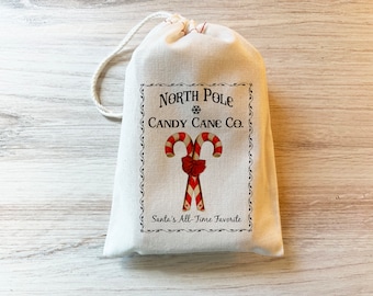 May include: A cream-colored drawstring pouch featuring a printed design of two red and white striped candy canes tied with a red bow. The text reads North Pole Candy Cane Co. and Santa's All-Time Favorite, enclosed within a decorative black border.