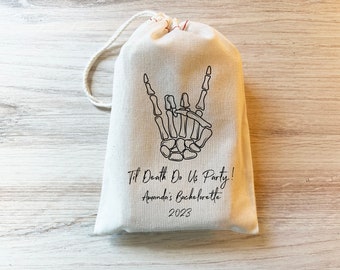 May include: A white cotton drawstring bag with a black skeleton hand making a rock and roll sign. The bag has the text "Til Death Do Us Party! Amanda's Bachelorette 2023" printed on it.
