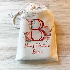 May include: A white fabric gift bag with a red letter "B" and a Santa Claus and reindeer illustration. The bag has the text "Merry Christmas Brian" printed on it.