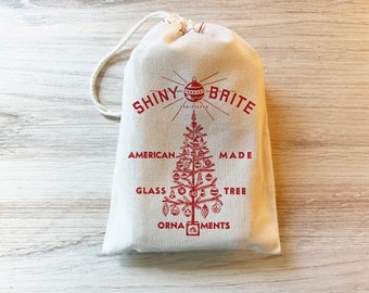 May include: A cream-colored fabric drawstring pouch featuring a red graphic print. The design includes the text SHINY BRITE above a stylized Christmas tree decorated with various ornaments, with the words AMERICAN MADE GLASS TREE ORNAMENTS.