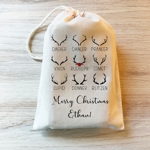 May include: A white fabric gift bag with a drawstring closure. The bag features a black and red design of reindeer antlers with the names of Santa's reindeer. The text "Merry Christmas Ethan!" is printed on the bag.