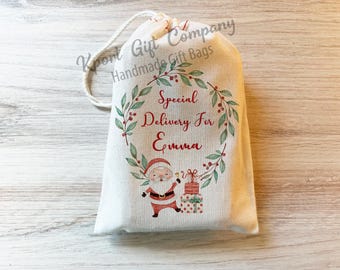 May include: A white fabric gift bag with a red and green wreath design. The bag has the text "Special Delivery For Emma" and a cartoon image of Santa Claus with a gift.