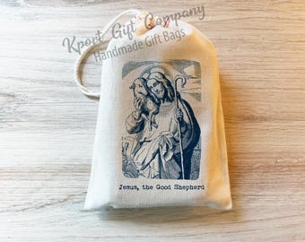 May include: A cream-colored fabric drawstring gift bag featuring a dark blue printed illustration of Jesus holding a lamb with a shepherd's staff. Below the image, the text reads: Jesus, the Good Shepherd. The bag has a white cord closure.