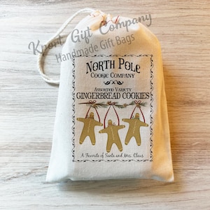 K&ouml;nnte beinhalten: Ein cremefarbener Geschenkbeutel mit Kordelzug und einem aufgedruckten Etikett mit der Aufschrift North Pole Cookie Company, Assorted Variety Gingerbread Cookies und A Favorite of Santa and Mrs. Claus sowie drei Lebkuchenm&auml;nnchen.
