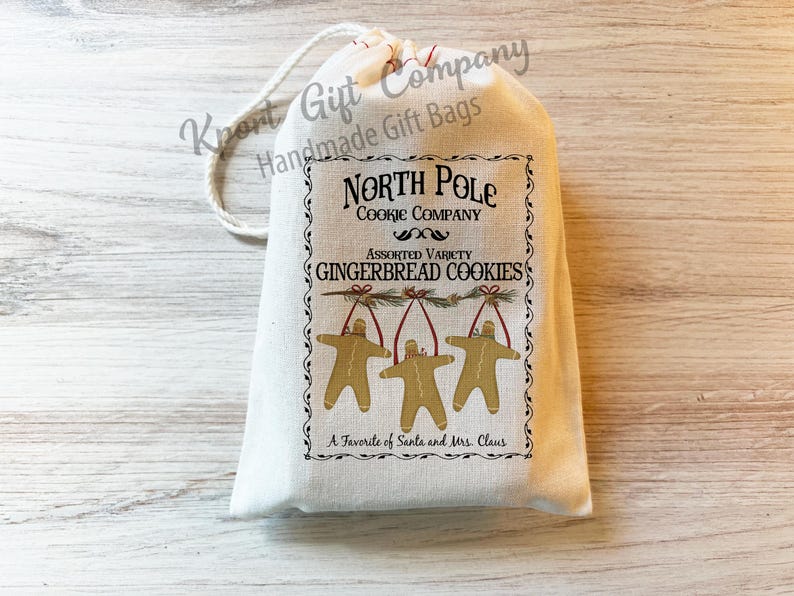 K&ouml;nnte beinhalten: Ein cremefarbener Geschenkbeutel mit Kordelzug und einem aufgedruckten Etikett mit der Aufschrift North Pole Cookie Company, Assorted Variety Gingerbread Cookies und A Favorite of Santa and Mrs. Claus sowie drei Lebkuchenm&auml;nnchen.