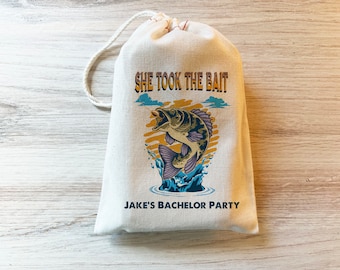 May include: A cream-colored canvas drawstring bag featuring a graphic of a jumping fish with the text "SHE TOOK THE BAIT" above it and "JAKE'S BACHELOR PARTY" printed in dark blue lettering below the fish illustration on the front of the bag.
