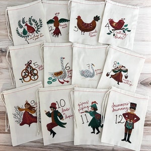 May include: A set of twelve off-white drawstring fabric bags, each featuring a unique illustration and number from one to twelve representing the Twelve Days of Christmas, with colorful printed graphics of birds, people, and objects in a folk art style.