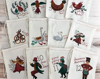 May include: A set of twelve off-white drawstring fabric bags, each featuring a unique illustration and number from one to twelve representing the Twelve Days of Christmas, with colorful printed graphics of birds, people, and objects in a folk art style.