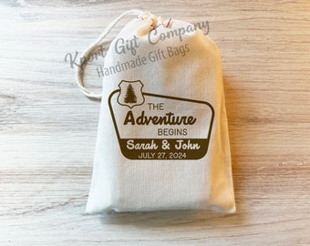 May include: A cream-colored fabric drawstring gift bag featuring a brown graphic of a pine tree inside a shield and a road sign shape. The text reads THE ADVENTURE BEGINS, Sarah & John, and JULY 27, 2024. The bag has a white cord closure.
