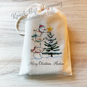 May include: A cream-colored drawstring gift bag featuring a printed illustration of three stacked snowmen decorating a snow-covered pine tree with a yellow star. The text Kport Gift Company Handmade Gift Bags and Merry Christmas Andrew is printed.