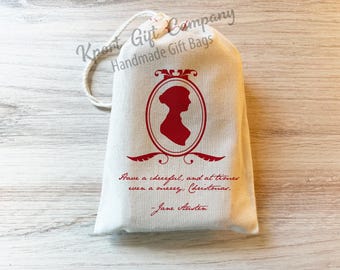 May include: A cream-colored fabric drawstring gift bag featuring a red silhouette of a person inside an oval frame. Below the frame, the text reads: Have a cheerful, and at times even a merry, Christmas. - Jane Austen. The bag has a white cord.