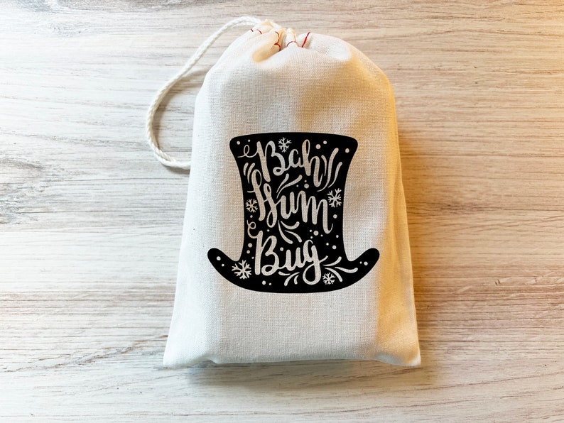 May include: A cream-colored drawstring pouch featuring a black graphic of a top hat. The hat contains the text "Bah Hum Bug" written in a stylized script font, surrounded by small snowflake and dot illustrations. The bag has a white cord closure.