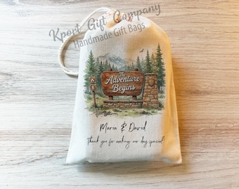 May include: A white fabric drawstring gift bag featuring a printed illustration of a wooden sign that reads The Adventure Begins, set against a forest and mountain backdrop. Below the graphic, the text reads Maria & David Thank you for making our day special!