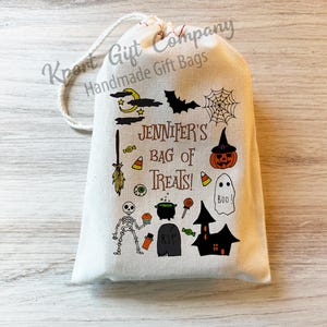 May include: A cream-colored Halloween-themed gift bag with a drawstring closure. The bag is decorated with various Halloween illustrations, including a bat, spiderweb, pumpkin, ghost, and skeleton. The text "Jennifer's Bag of Treats!" is printed on the front.