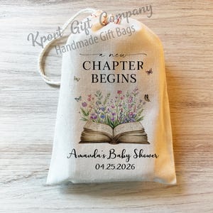 May include: A cream-colored fabric drawstring gift bag featuring a printed illustration of an open book with colorful wildflowers growing from the pages and small butterflies. The text reads A NEW CHAPTER BEGINS, Amanda's Baby Shower, and 04.25.2026.