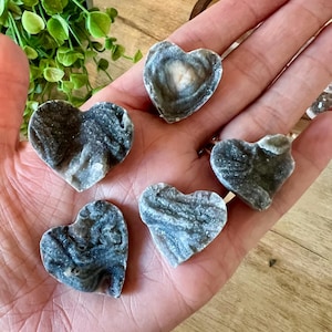 May include: A collection of six heart-shaped stones with a marbled appearance. The stones feature a mix of gray, white, and blue hues, with unique patterns. The stones are held in a hand, showcasing their natural beauty and texture.
