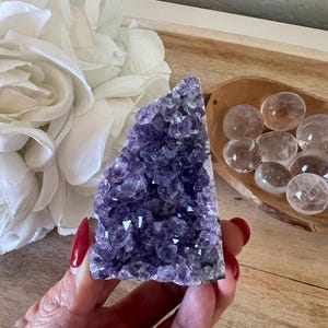 May include: A deep purple amethyst geode crystal cluster with a rough, textured surface. The crystal is held in a hand with red nail polish. Clear quartz spheres are in a wooden bowl in the background.