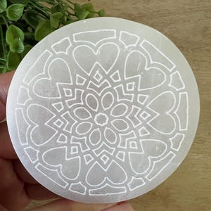 以下が含まれることがあります： ハートと幾何学模様がエッチングされた、丸い白いセレナイトクリスタルプレート。プレートは手で持たれており、彫刻されたパターンの複雑なディテールが示されています。クリスタルは滑らかで研磨された表面を持っています。