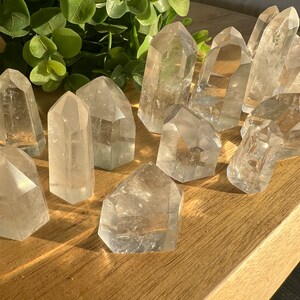 May include: A collection of clear quartz crystal points, also known as rock crystals, arranged on a wooden surface. The crystals are various sizes and shapes, with some having a pointed tip and others having a more rounded shape.