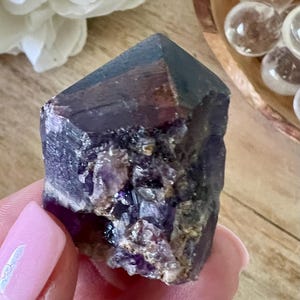 May include: A raw, faceted amethyst crystal with deep purple and brown hues. The crystal is held in a hand with a pink manicure, with clear quartz spheres and a white flower in the background.