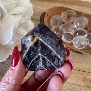 May include: A faceted, dark purple amethyst crystal with white veining is held in a hand with red nail polish. Clear quartz spheres sit in a wooden bowl, and a white flower is in the background.