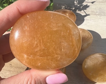 May include: A close-up of a smooth, orange-colored, polished stone. The stone is round and has a slightly irregular shape. It is being held in a hand with pink nail polish.
