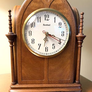 May include: A wooden mantel clock with a pointed arch top and two decorative side pillars. The white clock face features black gothic-style numerals, the word Sentinel, and ornate metal hands. The base is rectangular with a smooth wood grain finish.
