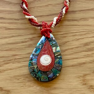 May include: A handcrafted necklace featuring a teardrop pendant and a twisted beaded chain. The pendant showcases a mosaic design with red, blue, and green elements, and a white spiral. The chain is made of red, white, and brown beads.