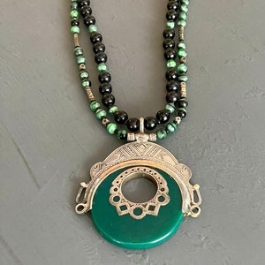 May include: A green and black beaded necklace with a large, round, green pendant. The pendant has a silver frame with intricate designs.