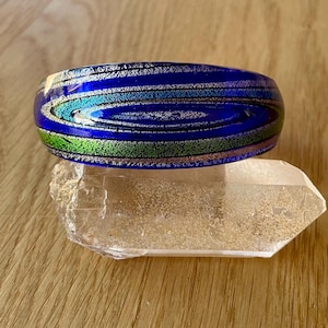 May include: A vibrant blue glass bangle bracelet with swirling patterns of teal, green, and silver. The bracelet rests on a clear quartz crystal, set against a wooden surface.
