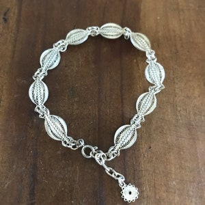 May include: A silver bracelet with a delicate filigree design. The bracelet is made of small, oval-shaped links that are connected together to form a chain. The chain is secured with a clasp.