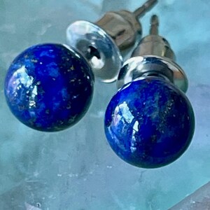 May include: A pair of stud earrings featuring deep blue, spherical stones with flecks of gold. The earrings have a silver post and backing. The stones have a polished, reflective surface, and the earrings are set against a light blue background.