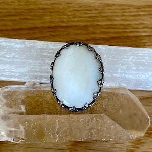 May include: Oval-shaped ring featuring a creamy white stone set in a dark, ornate metal frame. The ring's bezel has a detailed, textured design. The ring is displayed on a light-colored surface, with a crystal and a white stone in the background.