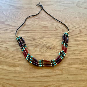May include: A three-strand necklace with a black cord. The necklace features dark red, turquoise, and gold-colored beads. The beads are arranged in a repeating pattern. The necklace is displayed on a wooden surface.