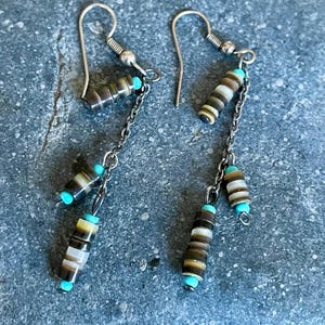 May include: A pair of dangle earrings with silver-tone hooks and chains. The earrings feature cylindrical beads in shades of brown, tan, and white, accented with small turquoise beads. The earrings are displayed on a textured gray surface.
