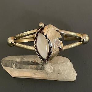 May include: A silver bracelet with a white mother of pearl stone and a leaf design. The bracelet is resting on a clear quartz crystal.