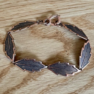 May include: A copper-toned bracelet featuring a series of stylized leaf-shaped links. Each leaf has a dark, textured interior and a copper-colored outline. The bracelet is connected with a matching clasp, and is displayed on a wooden surface.