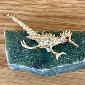May include: A gold-colored roadrunner bird brooch with a red gemstone eye, resting on a green stone. The brooch has detailed feather and body features. The bird's beak is pointed, and its tail is extended.