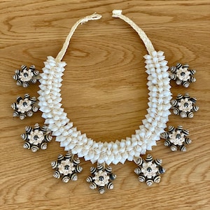May include: A handcrafted necklace featuring a white shell design with decorative black and white accents. The necklace is made of small, white shells and has a woven cord. The necklace has a unique, floral-like design.