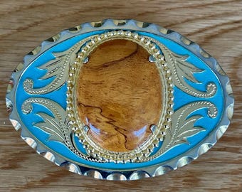May include: Oval-shaped belt buckle with a light blue enamel background and a gold-colored filigree design. The center features a polished, oval wooden inlay. The buckle has a silver-colored scalloped edge.